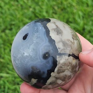 May include: A polished stone sphere featuring a distinct color split between a translucent blue-grey section with dark inclusions and an opaque, mottled beige and brown area. The surface is smooth and rounded, showcasing natural mineral patterns.
