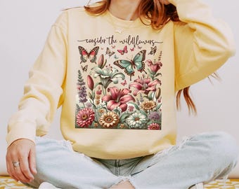 May include: Pale yellow sweatshirt featuring a floral design with butterflies and the text "consider the wildflowers." The graphic includes pink, red, and teal butterflies, and various flowers. The sweatshirt is long-sleeved.