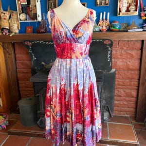 May include: A sleeveless dress featuring a vibrant landscape print with deep red trees, purple foliage, and light blue sky tones. The dress has a V-neckline, a fitted bodice, and a pleated skirt that falls to the knee, creating a full silhouette.