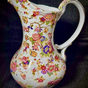 May include: A white porcelain pitcher with a floral pattern in shades of pink, red, yellow, blue, and green. The pitcher has a wide mouth and a curved handle.
