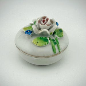 May include: A small, white ceramic trinket box with a lid, adorned with a delicate, hand-painted rose on top. The rose features soft pink and white petals, surrounded by green leaves and small blue flowers. The box has a gold trim.