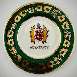 May include: A white ceramic plate with a green and gold border. The plate features a coat of arms with a red shield and gold accents. The text "MC INERNEY" is printed below the coat of arms.