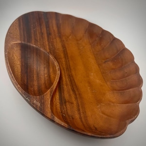 May include: A wooden serving dish with a natural wood grain finish. The dish has an oval shape with a smaller, attached compartment on the left side. The right side has a scalloped edge. The dish is made of a warm brown wood.