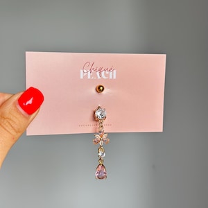 May include: A rose gold belly button ring with a dangling design. The top has a small gold ball. The dangling section includes a clear gem, a flower with pink and clear gems, another clear gem, and a pink teardrop gem. The text "Chique Peach" is on the pink card.