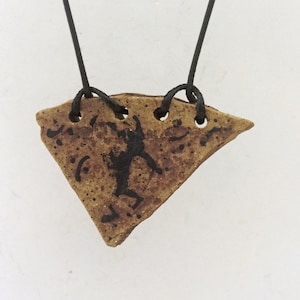 May include: A pendant necklace featuring a triangular, tan-colored ceramic pendant with a black figure design. The pendant is suspended from a black cord. The necklace has a rustic, handmade aesthetic.