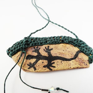 May include: A brown ceramic pendant with a black lizard design, accented with a dark green crocheted trim and a dark green cord with crystal beads.
