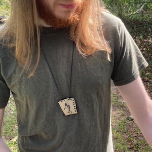 May include: A person wearing a green shirt with a necklace featuring a brown pendant with a black and white drawing of a person dancing. The pendant is attached to a black cord.