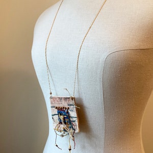 May include: A square-shaped pendant necklace with a woven fabric design and brown, blue, and white accents. The pendant is attached to a long, thin, brown cord.