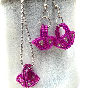 May include: A beaded jewelry set featuring a necklace and earrings. The necklace has a silver chain with a small, pink beaded basket pendant. The earrings are also pink beaded baskets, hanging from silver hooks. The jewelry is displayed against a light blue and white background.