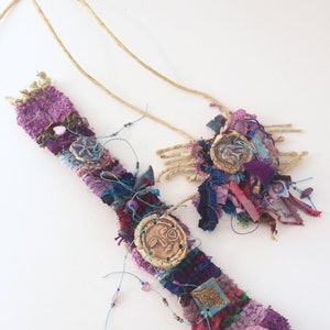 May include: A handmade necklace and bracelet set with a woven fabric base and colorful embellishments. The necklace features a large, round, ceramic pendant with a face design. The bracelet has a smaller, round, ceramic button with a floral design.