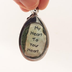 May include: A silver oval pendant with a clear glass front. The glass contains a piece of paper with the words "My Heart To Your Heart" written on it. The paper is surrounded by green moss.