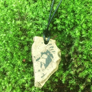 May include: A ceramic pendant with a black and white design hangs from a black cord. The pendant is shaped like a teardrop and is resting on a bed of green moss.