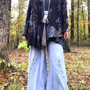 May include: A dark floral print jacket with a gray top and light blue wide-leg pants. The jacket features a pattern of blue, brown, and gray leaves. The pants have button details along the side seams. A silver necklace with a unique pendant is also visible.
