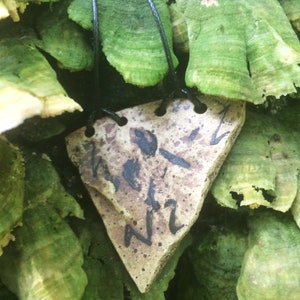May include: A triangular ceramic pendant with black ink markings, including the letters "NZ" and "HOT", hanging from a black cord against a backdrop of green and white lichen.