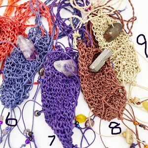 May include: Close-up of several handmade macrame pouches in various colors, including red, blue, purple, brown, and beige. Each pouch contains a crystal or gemstone. The pouches are strung with cords and adorned with beads.