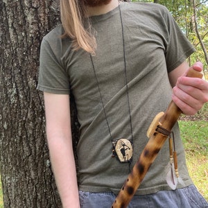 May include: A person wearing a green t-shirt and grey pants is holding a wooden flute with a leather strap and a small, round, beaded pendant. The flute has a feather attached to it.