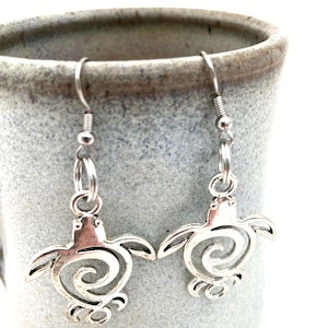 May include: A pair of silver-colored earrings featuring a sea turtle design. The earrings have a hook closure and a detailed turtle pendant with a spiral design on the shell. The earrings are displayed against a neutral background.