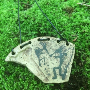 May include: A ceramic pendant with a black and white design of two figures. The pendant is attached to a black cord and is sitting on a bed of green moss.