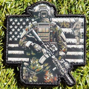 May include: A black and white American flag patch with a soldier in camouflage holding a rifle. The soldier is standing in front of the flag.