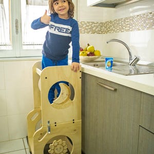 Può includere: Un bambino sta in piedi su una torre di apprendimento Montessori in legno con un design di luna e sole. La torre si trova in un ambiente da cucina con un lavandino e armadi.