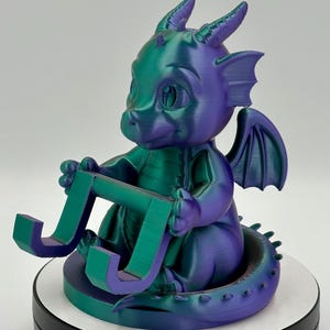 May include: A teal and purple 3D-printed dragon figure holding the letter "J". The dragon has horns, wings, and a tail. The figurine sits on a white and black circular base. The colours are a mix of teal and purple.