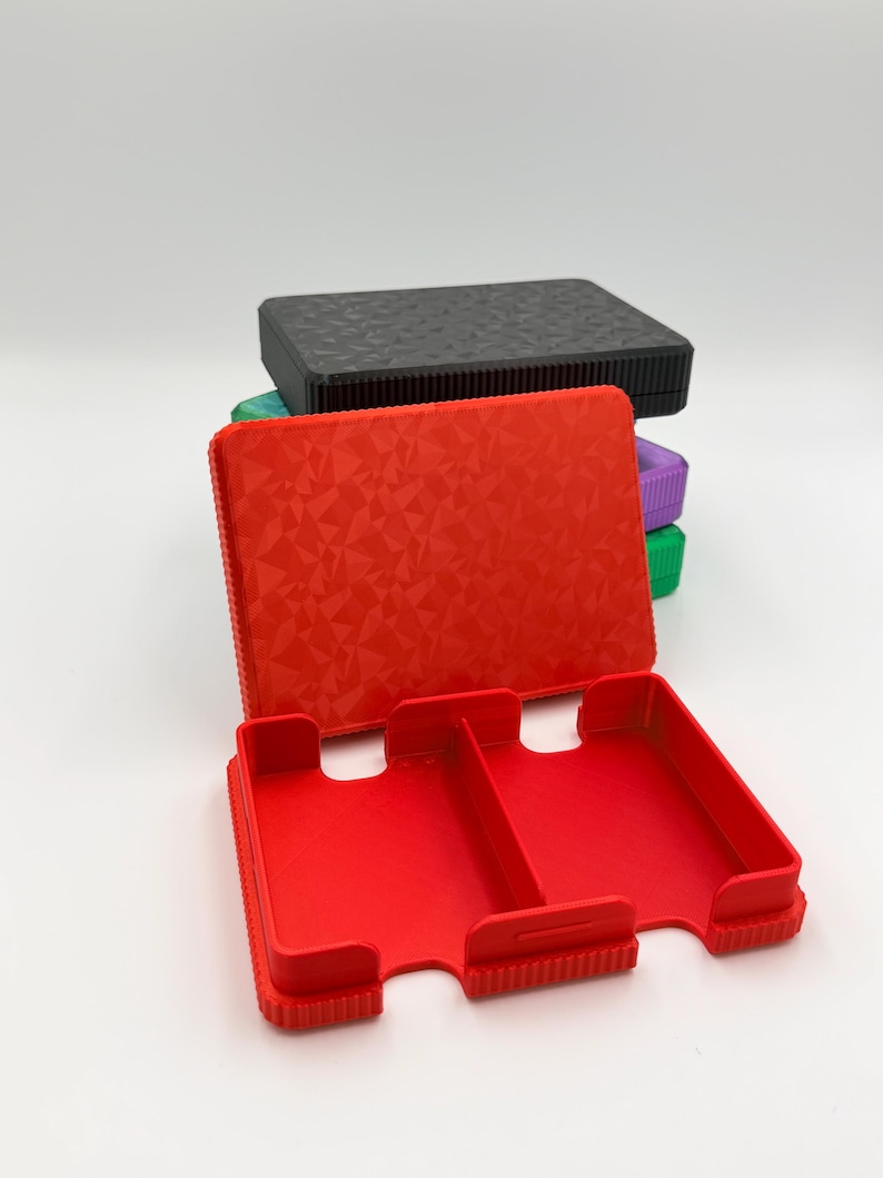 May include: A red, open, rectangular case with two compartments, featuring a textured geometric pattern on the lid. A stack of similar cases in black, green, and purple are visible in the background. The cases appear to be made of plastic.