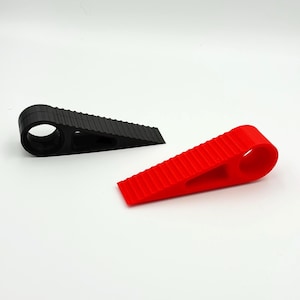 Puede incluir: Dos topes de puerta, uno negro y otro rojo, sobre un fondo blanco. Cada tope de puerta tiene forma de cuña con una superficie estriada y una abertura circular. Están diseñados para evitar que las puertas se cierren de golpe.