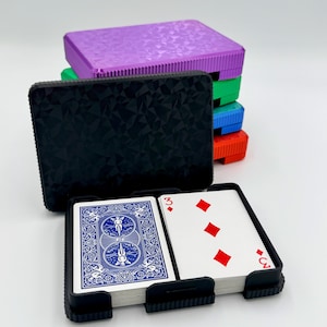 May include: A black playing card holder with a deck of cards featuring a blue back design and red diamond suits. A stack of colorful, textured card cases in purple, green, blue, and red are behind the holder.