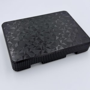 May include: A black, rectangular object with a geometric pattern on the top surface. The sides have a ridged texture, and the object has small feet. The overall design is modern and minimalist.