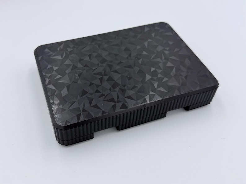 May include: A black, rectangular object with a geometric pattern on the top surface. The sides have a ridged texture, and the object has small feet. The overall design is modern and minimalist.