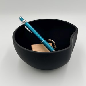 Puede incluir: Un cuenco negro con acabado mate y un diseño curvo único. En el interior, un bolígrafo turquesa, un pequeño bloque de madera y un llavero dorado son visibles. El cuenco se utiliza probablemente para guardar objetos pequeños como llaves o bolígrafos.