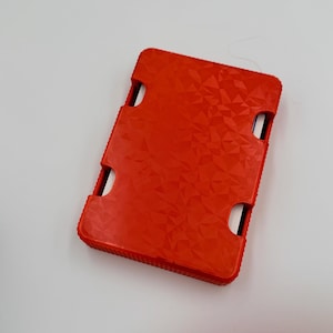 May include: A bright red rectangular case with a textured geometric pattern. The case has four curved cutouts and a ridged edge. The case is likely designed to hold a small electronic device.