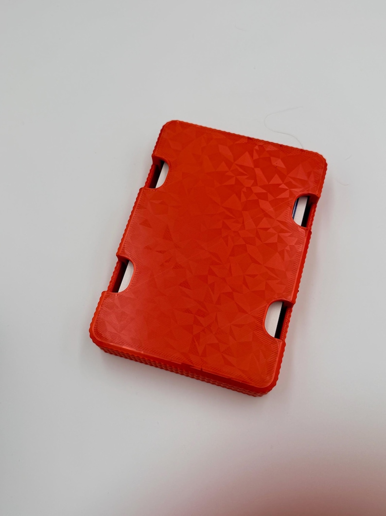 May include: A bright red rectangular case with a textured geometric pattern. The case has four curved cutouts and a ridged edge. The case is likely designed to hold a small electronic device.