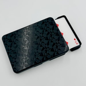 May include: Black playing card case with a geometric pattern. A playing card with the three of diamonds is partially visible. The case is rectangular and has a textured surface.