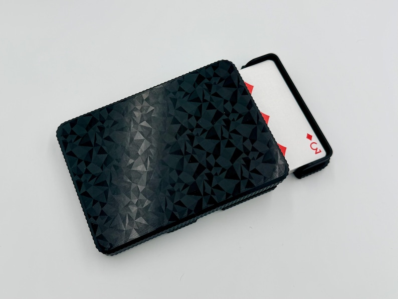 May include: Black playing card case with a geometric pattern. A playing card with the three of diamonds is partially visible. The case is rectangular and has a textured surface.