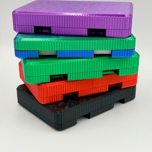 May include: A stack of colorful, textured plastic boxes in purple, green, red, and black. Each box has a rectangular shape with a ridged surface and a recessed area on the sides. The boxes are stacked on top of each other.