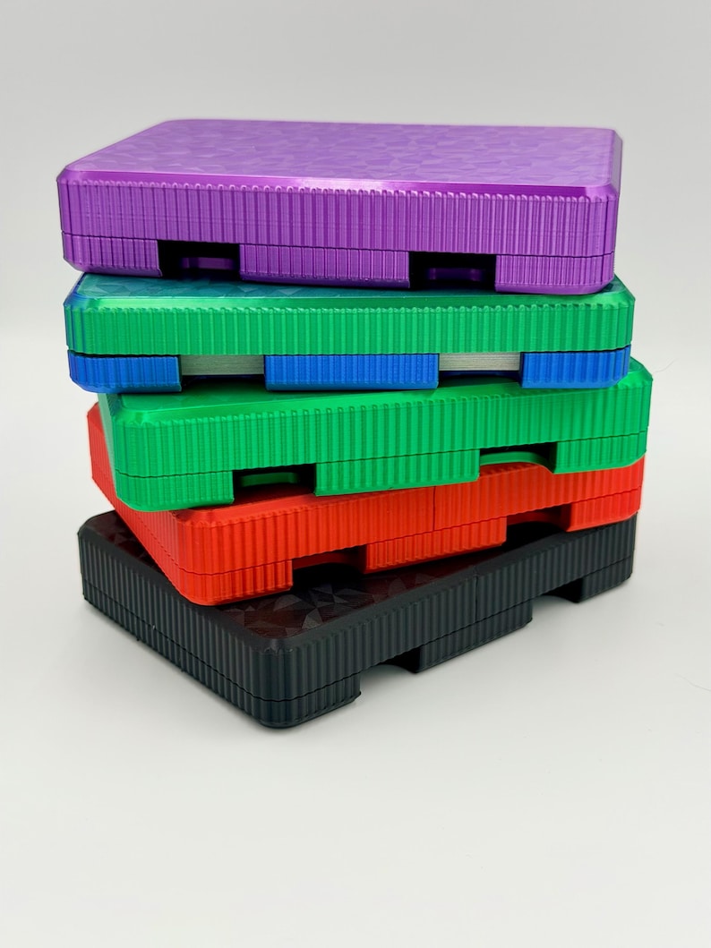 May include: A stack of colorful, textured plastic boxes in purple, green, red, and black. Each box has a rectangular shape with a ridged surface and a recessed area on the sides. The boxes are stacked on top of each other.