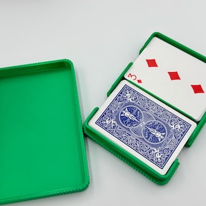 May include: A green plastic playing card case with a deck of cards inside. The cards have a blue back design and the face-up card shows the three of diamonds. The case is open, revealing the cards.