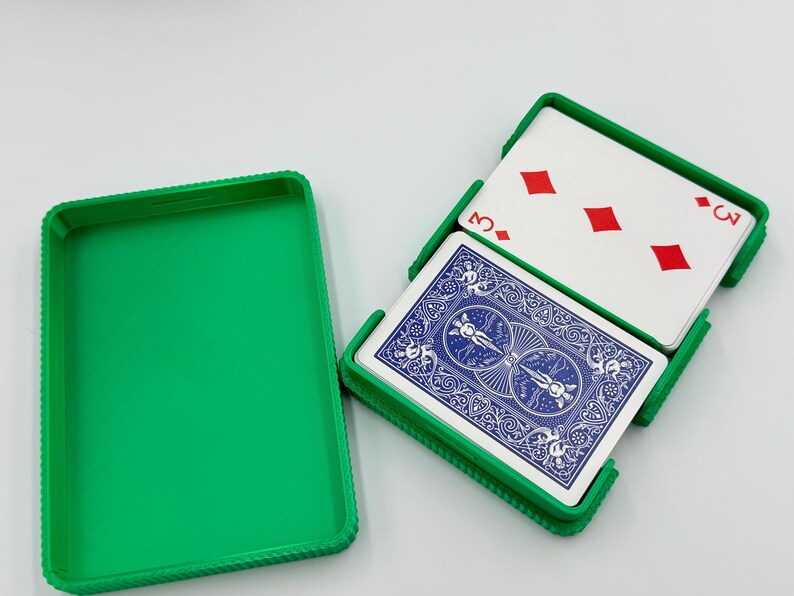 May include: A green plastic playing card case with a deck of cards inside. The cards have a blue back design and the face-up card shows the three of diamonds. The case is open, revealing the cards.
