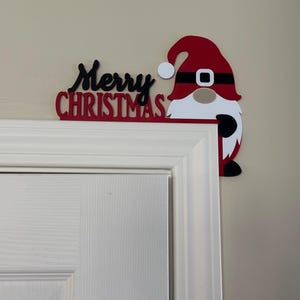 Close-up view of the Christmas gnome sign resting on the upper right corner of a white door frame against a beige wall.