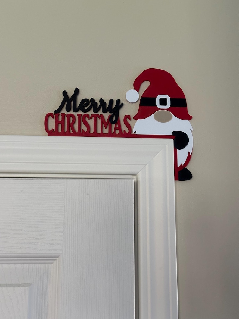 Close-up view of the Christmas gnome sign resting on the upper right corner of a white door frame against a beige wall.
