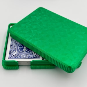 May include: A bright green playing card case, partially open, revealing a deck of cards with blue backs. The case has a textured geometric pattern on the lid and a ribbed edge. The case is designed to hold a standard deck of cards.