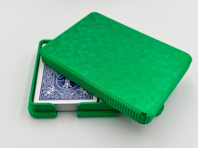 May include: A bright green playing card case, partially open, revealing a deck of cards with blue backs. The case has a textured geometric pattern on the lid and a ribbed edge. The case is designed to hold a standard deck of cards.