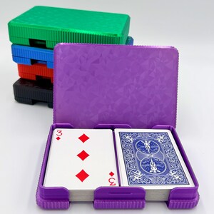 May include: A purple playing card case with a deck of cards inside, showing the three of diamonds. A stack of colorful card cases in green, blue, red, and black are behind the open case. The card case has a textured geometric pattern.