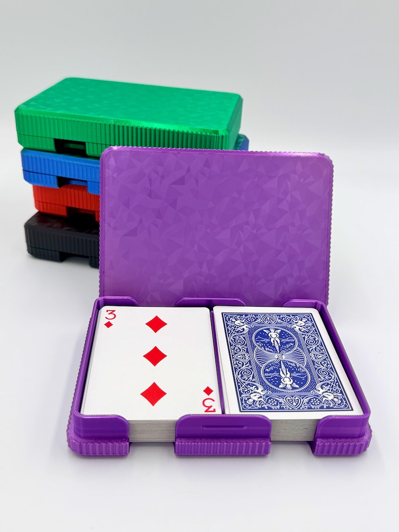 May include: A purple playing card case with a deck of cards inside, showing the three of diamonds. A stack of colorful card cases in green, blue, red, and black are behind the open case. The card case has a textured geometric pattern.