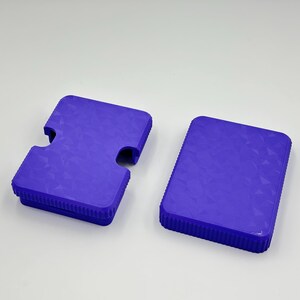May include: Two blue plastic cases with textured surfaces. One case has a unique cutout shape, while the other is rectangular. Both cases have a ridged border. The cases are likely for storing or protecting items.