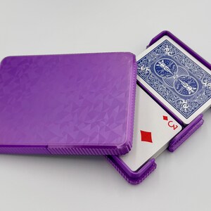 May include: A purple playing card case with a geometric pattern, partially open to reveal a deck of cards. The cards have a blue back design and the three of diamonds is visible.