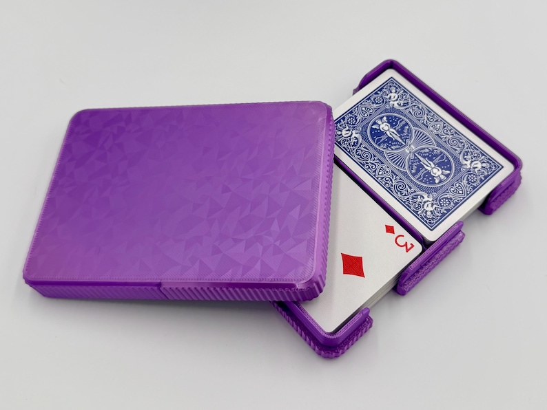 May include: A purple playing card case with a geometric pattern, partially open to reveal a deck of cards. The cards have a blue back design and the three of diamonds is visible.