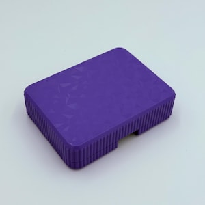 May include: A rectangular, purple box with a textured geometric pattern on the top surface. The sides of the box have a ribbed design. The box is set against a white background.