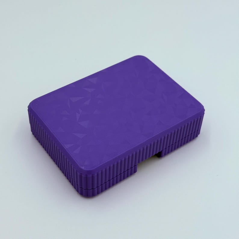 May include: A rectangular, purple box with a textured geometric pattern on the top surface. The sides of the box have a ribbed design. The box is set against a white background.